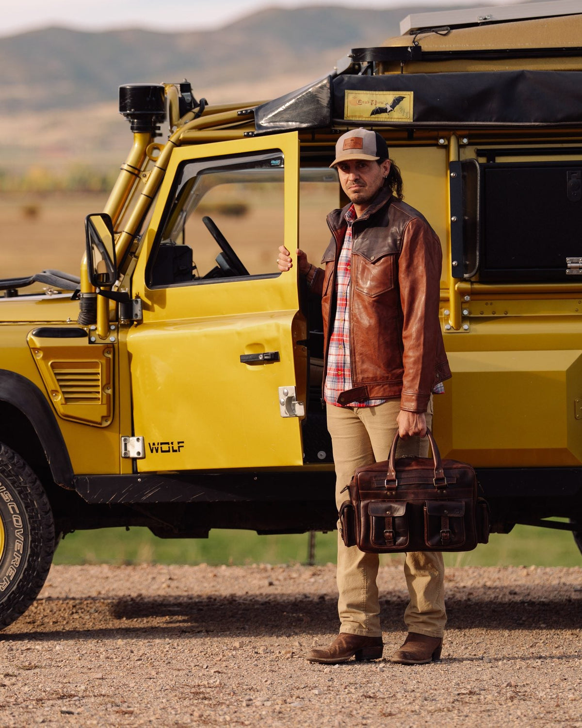 Man in a two-tone Western leather jacket and plaid flannel holding a leather pilot bag beside a yellow Land Rover