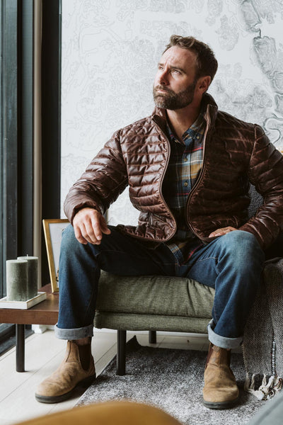 Casual look: man in a brown quilted jacket and jeans, sitting indoors with a contemplative expression.