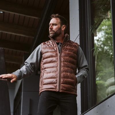 Man sporting a brown quilted vest, walking in a scenic outdoor setting, looking reflective.