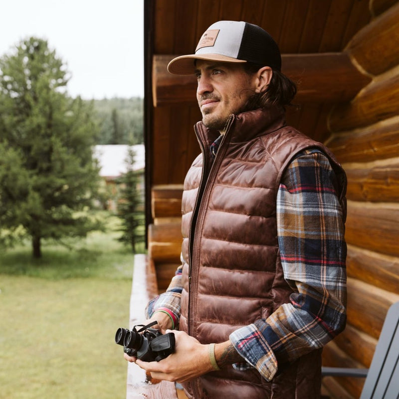 Outdoorsman in a brown puffer vest, walking and gazing into the distance with binoculars in hand.