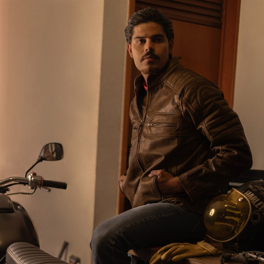 Man in the Cafe Racer Leather Jacket in Distressed Brown seated by his motorcycle in a garage, jacket front in view.
