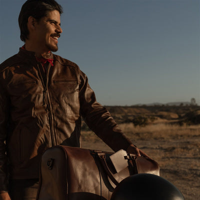 Man in the Cafe Racer Leather Jacket in Distressed Brown beside a motorcycle loaded with a leather duffle.