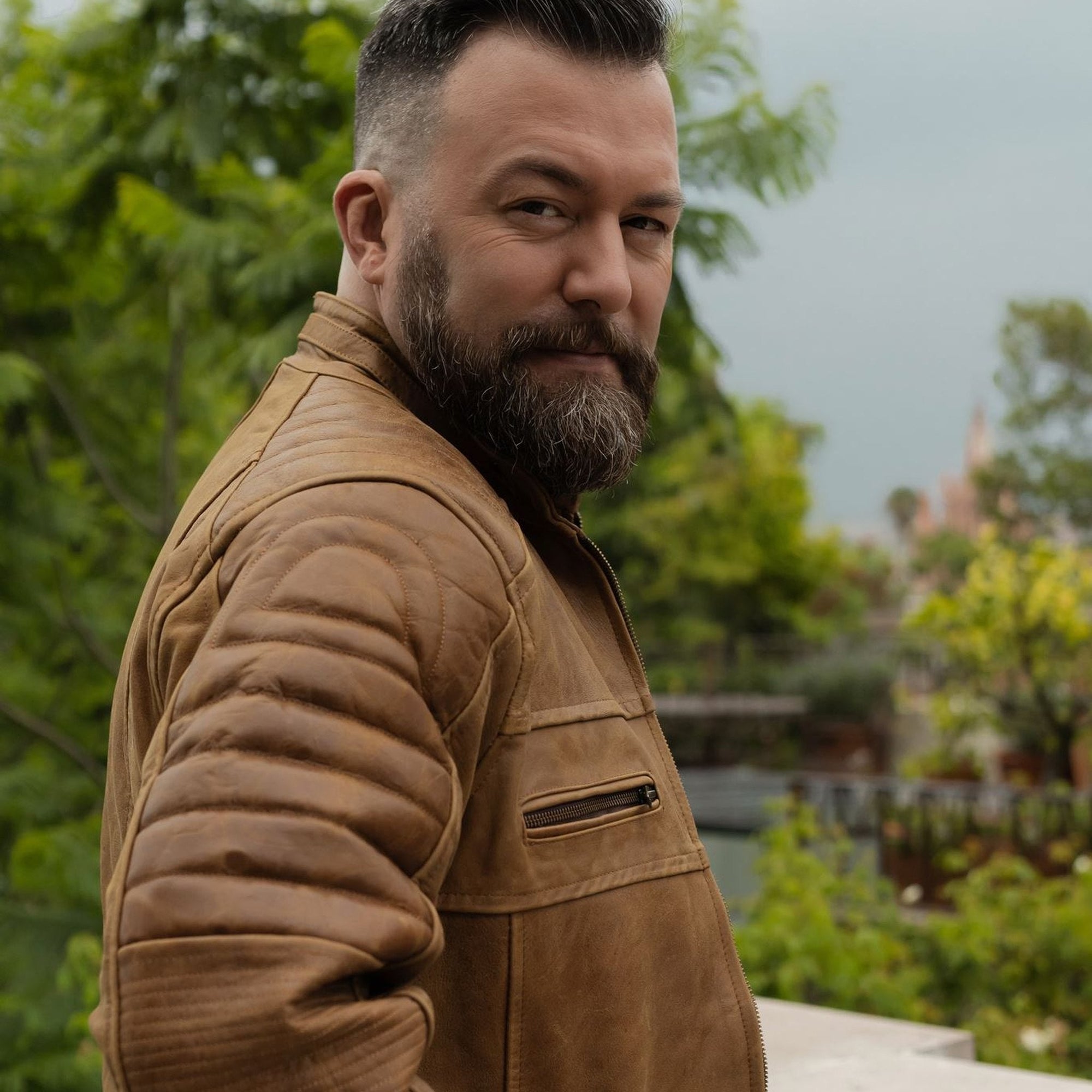 Man in the Cafe Racer Leather Jacket in Distressed Tan, side view showing the ribbed shoulders, on a garden balcony.