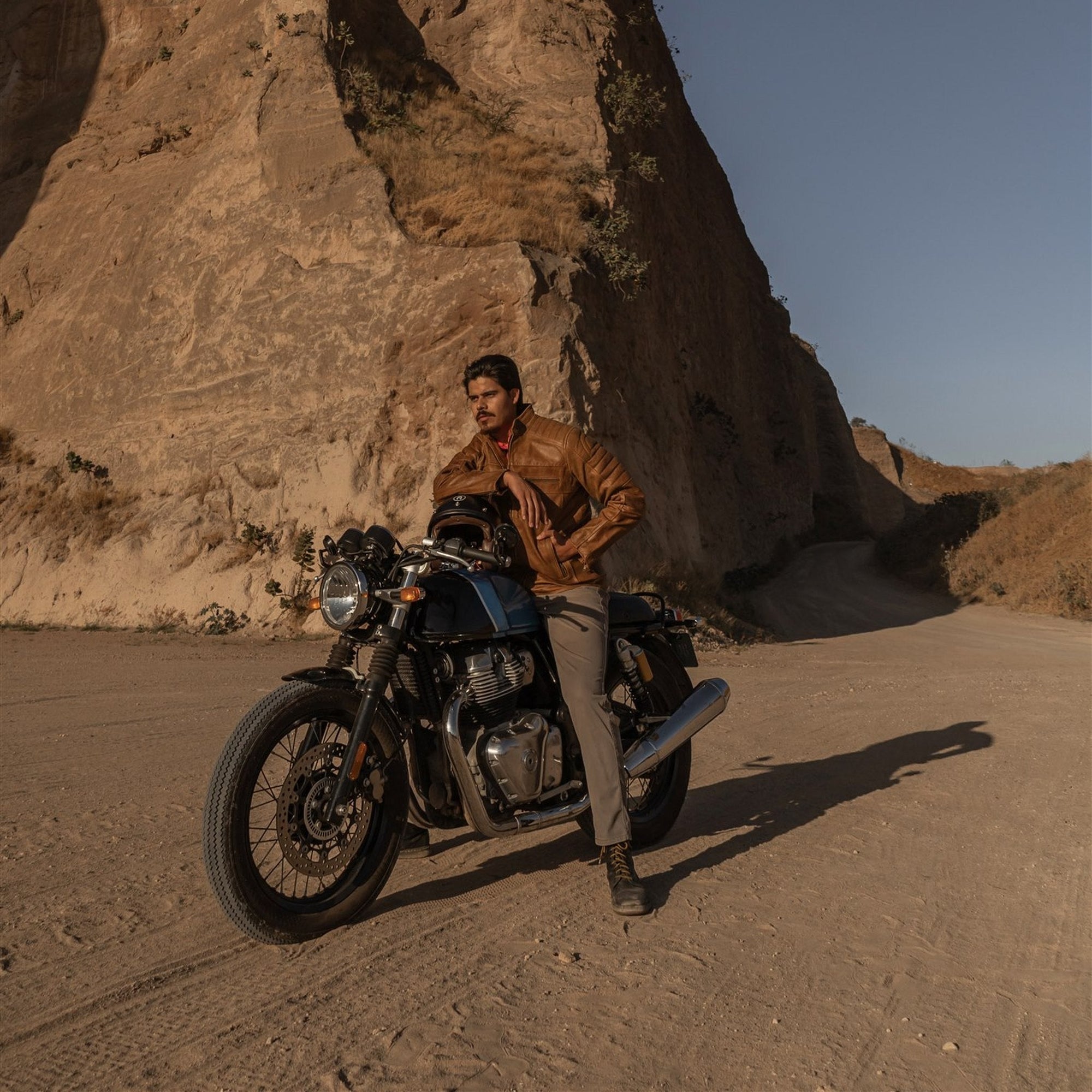 Rider in the Cafe Racer Leather Jacket in Distressed Tan with his motorcycle below a desert cliff.