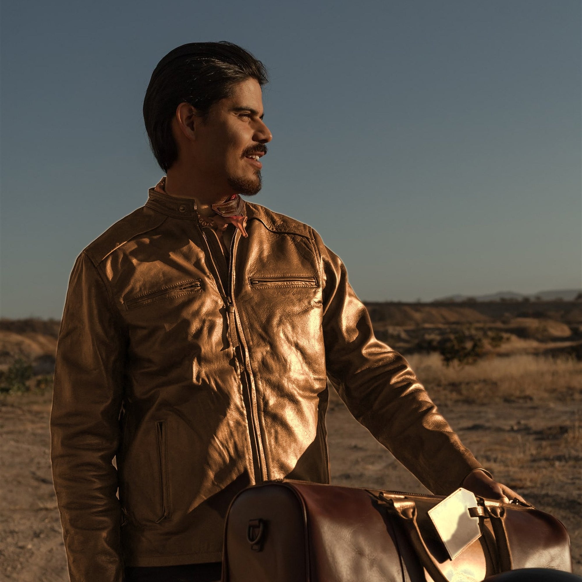 Man in the Cafe Racer Leather Jacket in Distressed Tan beside a motorcycle loaded with a leather duffle.