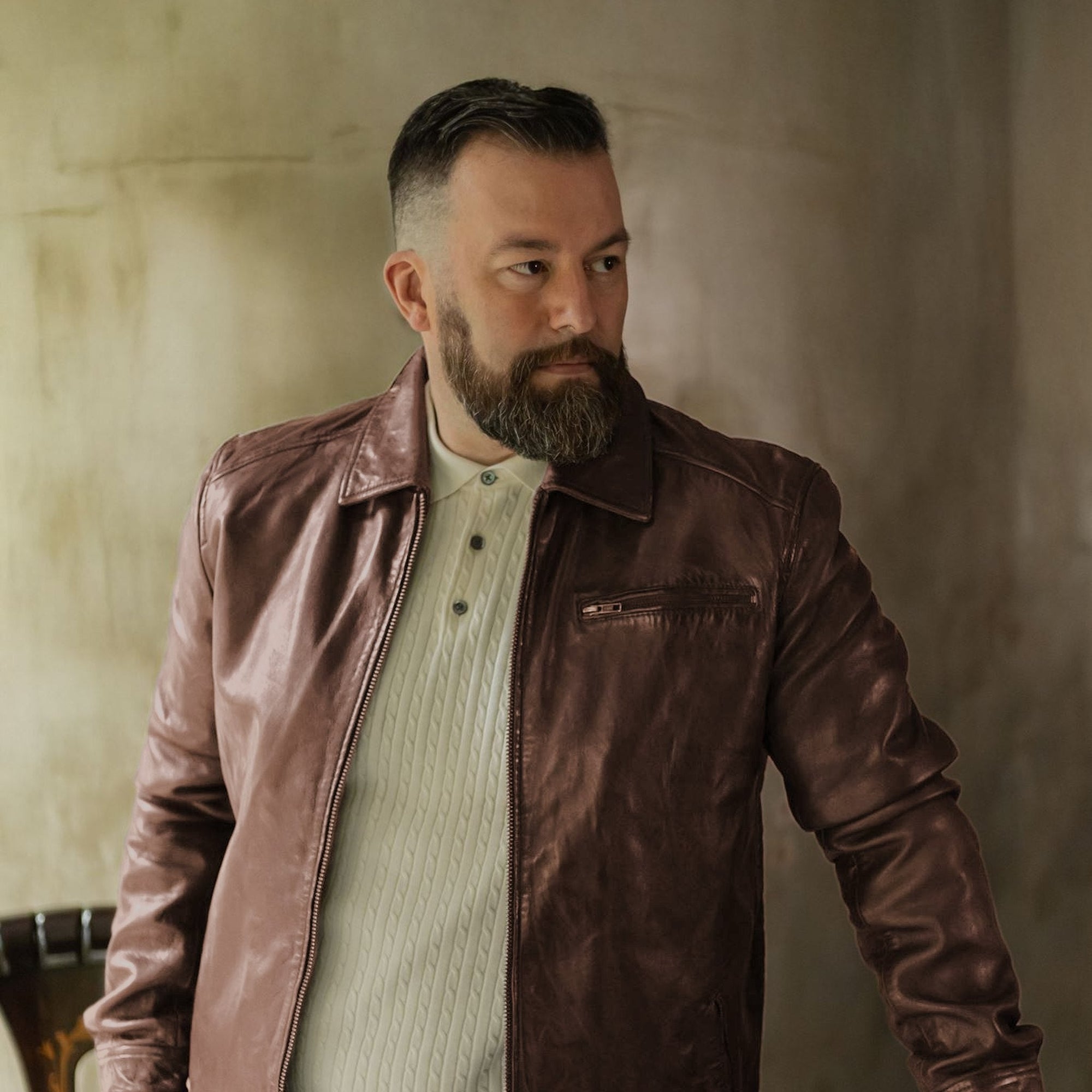Man in the Glenwood Leather Driving Jacket in Mahogany Brown over a cream henley, standing in a sage-walled room.