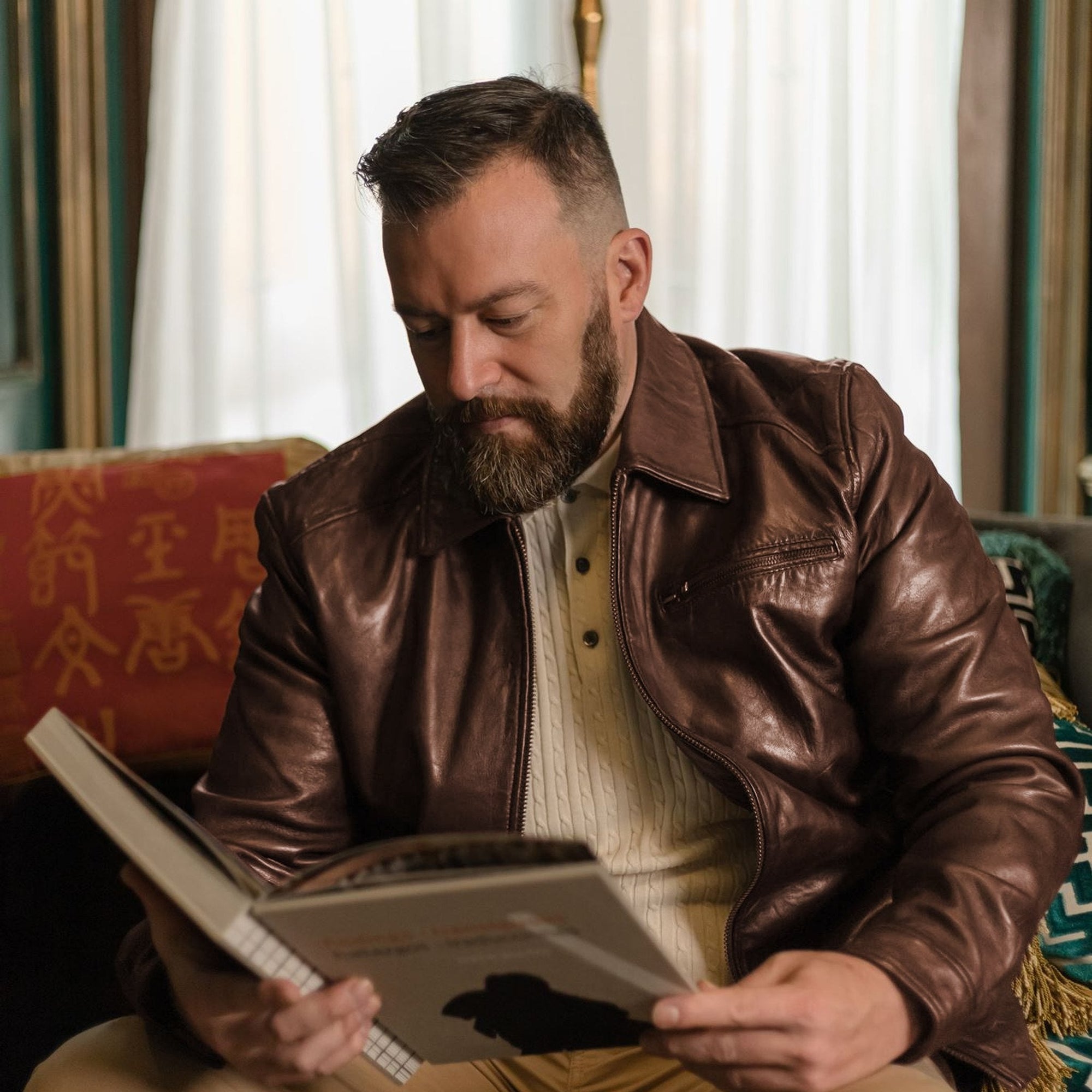 Man in the Glenwood Leather Driving Jacket in Mahogany Brown reading a large book on a red sofa by a window.