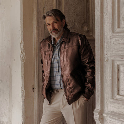 Man in the Glenwood Leather Driving Jacket in Mahogany Brown and a plaid shirt, leaning in a weathered wooden doorway.