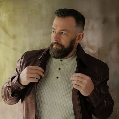 Man holding the lapels of the Glenwood Leather Driving Jacket in Mahogany Brown, worn open over a cream knit polo against a sage wall.
