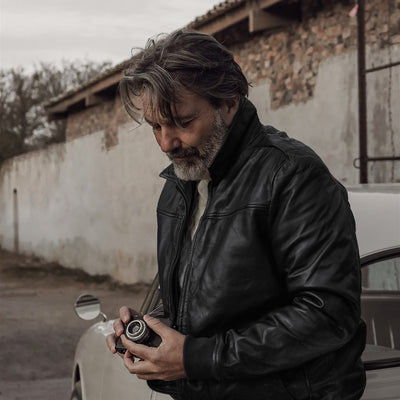 Man in the Glenwood Leather Driving Jacket in Vintage Black checking a film camera beside a white vintage car.