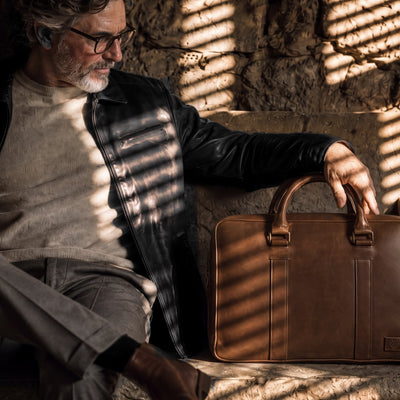 Man in the Glenwood Leather Driving Jacket in Vintage Black seated on a stone bench in slatted sunlight, a leather briefcase beside him.