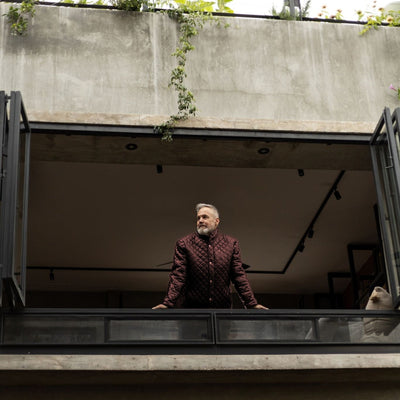 Man in the Highlands Quilted Leather Jacket in Mahogany Brown on an open-air balcony.
