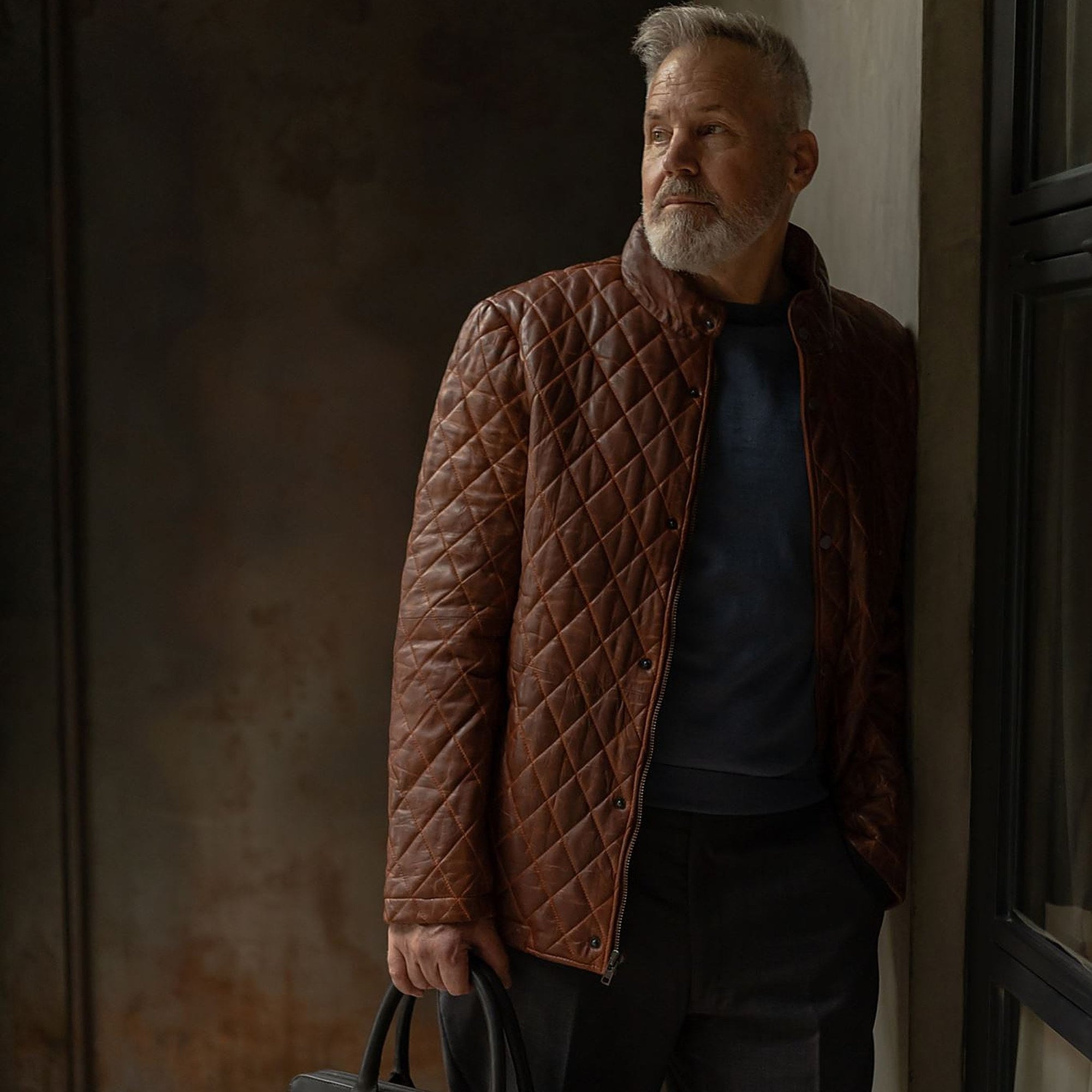 Man in the Highlands Quilted Leather Jacket in Whiskey Brown in a doorway, carrying a black leather briefcase.