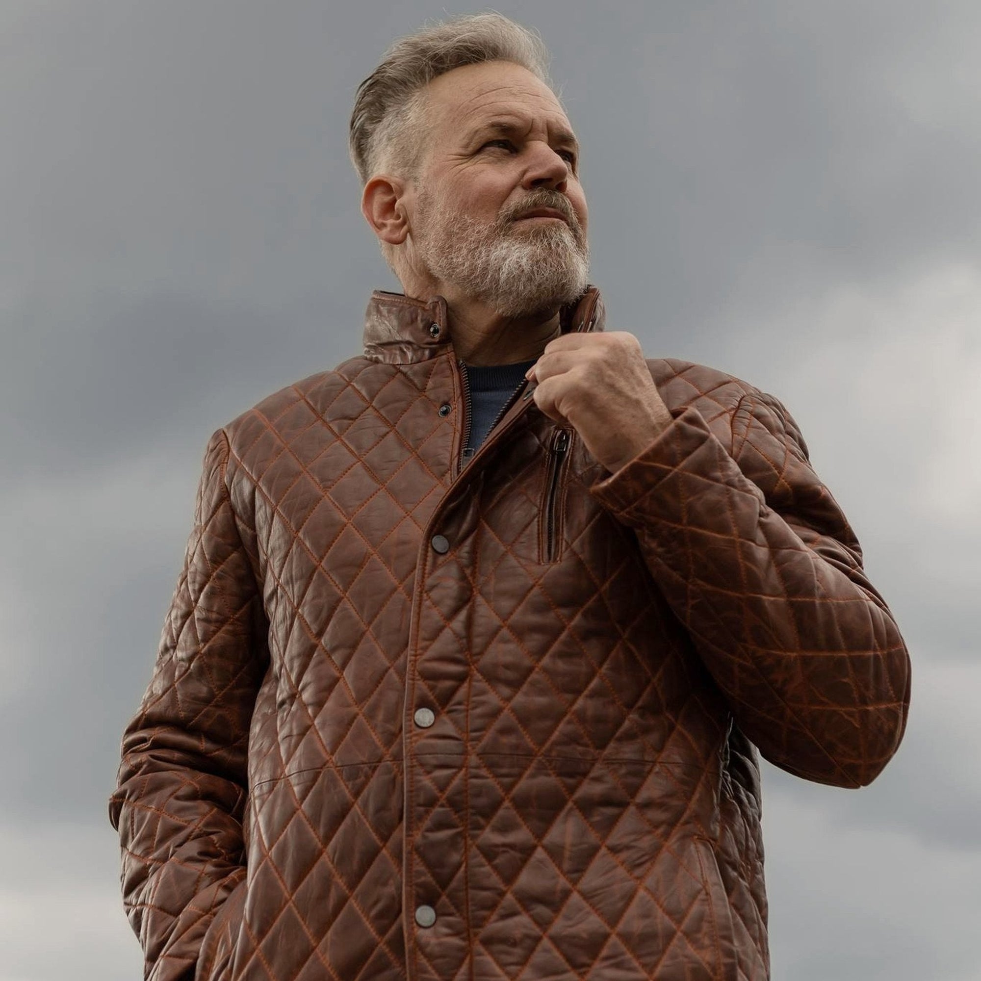 Man in the Highlands Quilted Leather Jacket in Whiskey Brown on a rooftop under a stormy sky.