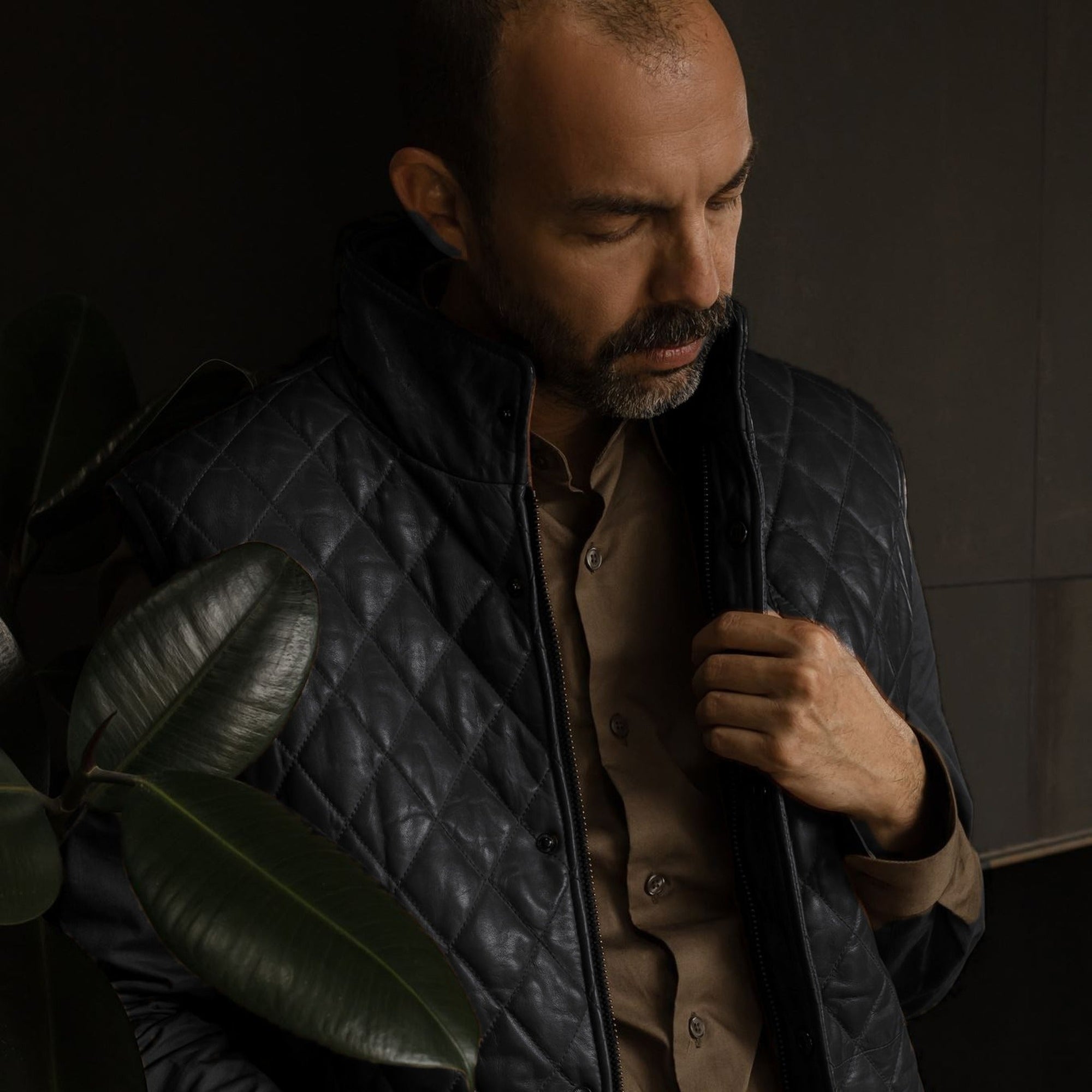Man in the Highlands Quilted Leather Vest in Black in a dark room with plants.