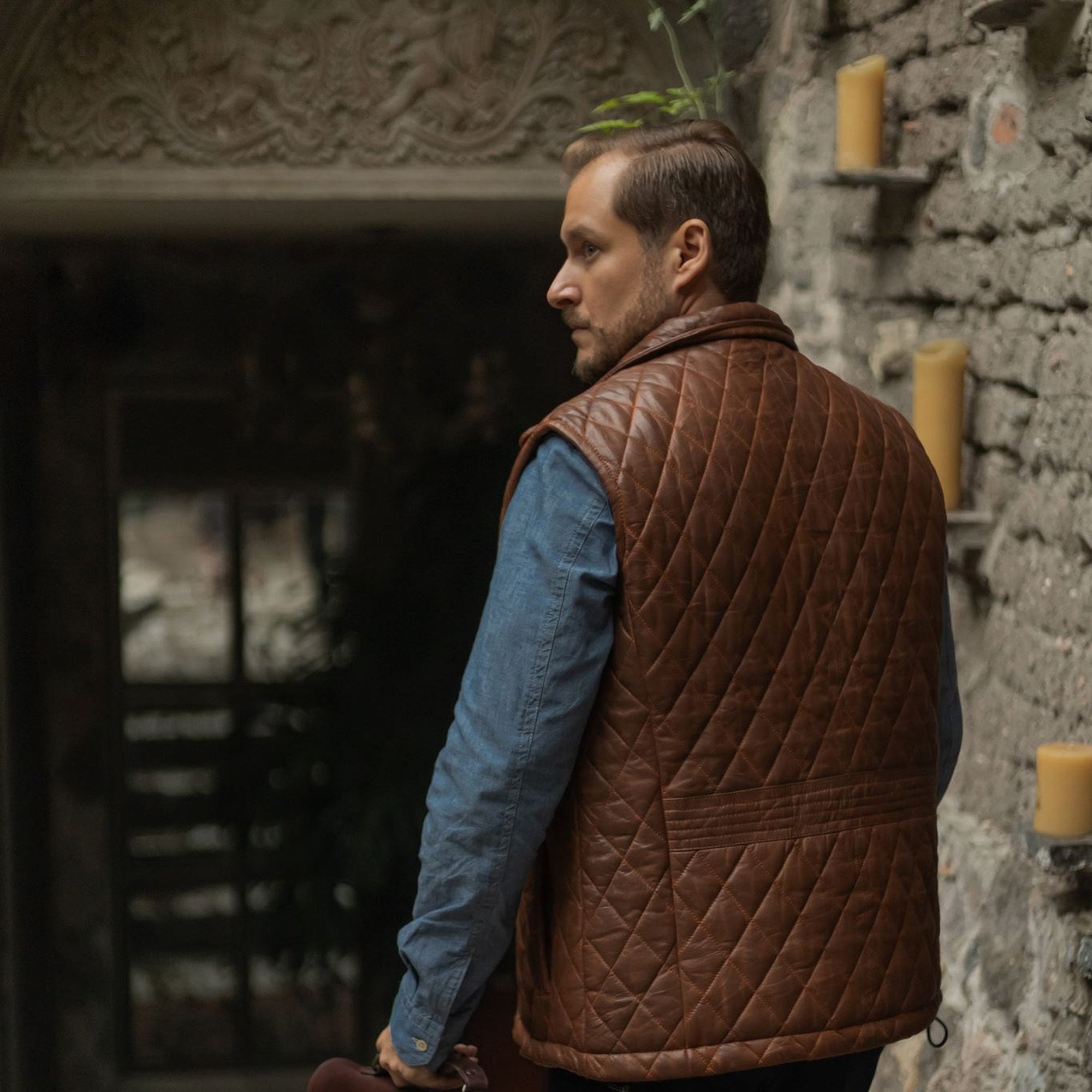 Back view of the Highlands Quilted Leather Vest in Whiskey Brown under a stone archway.