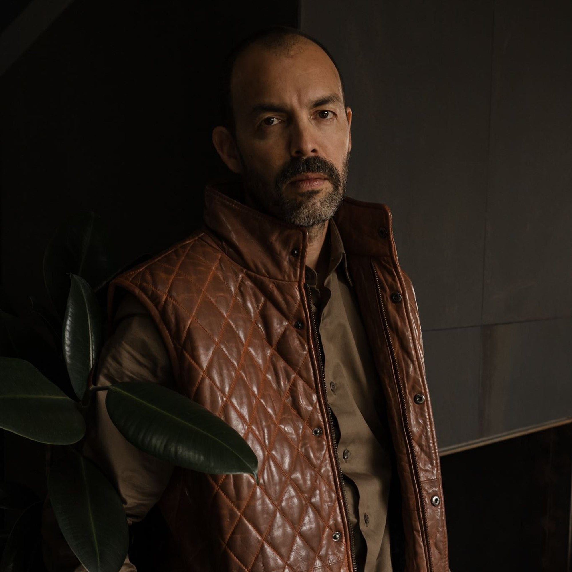 Man adjusting the collar of the Highlands Quilted Leather Vest in Whiskey Brown in a dark room with plants.