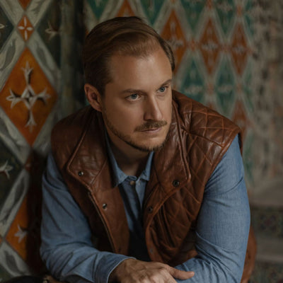 Man seated in a tiled room in the Highlands Quilted Leather Vest in Whiskey Brown over a blue shirt.