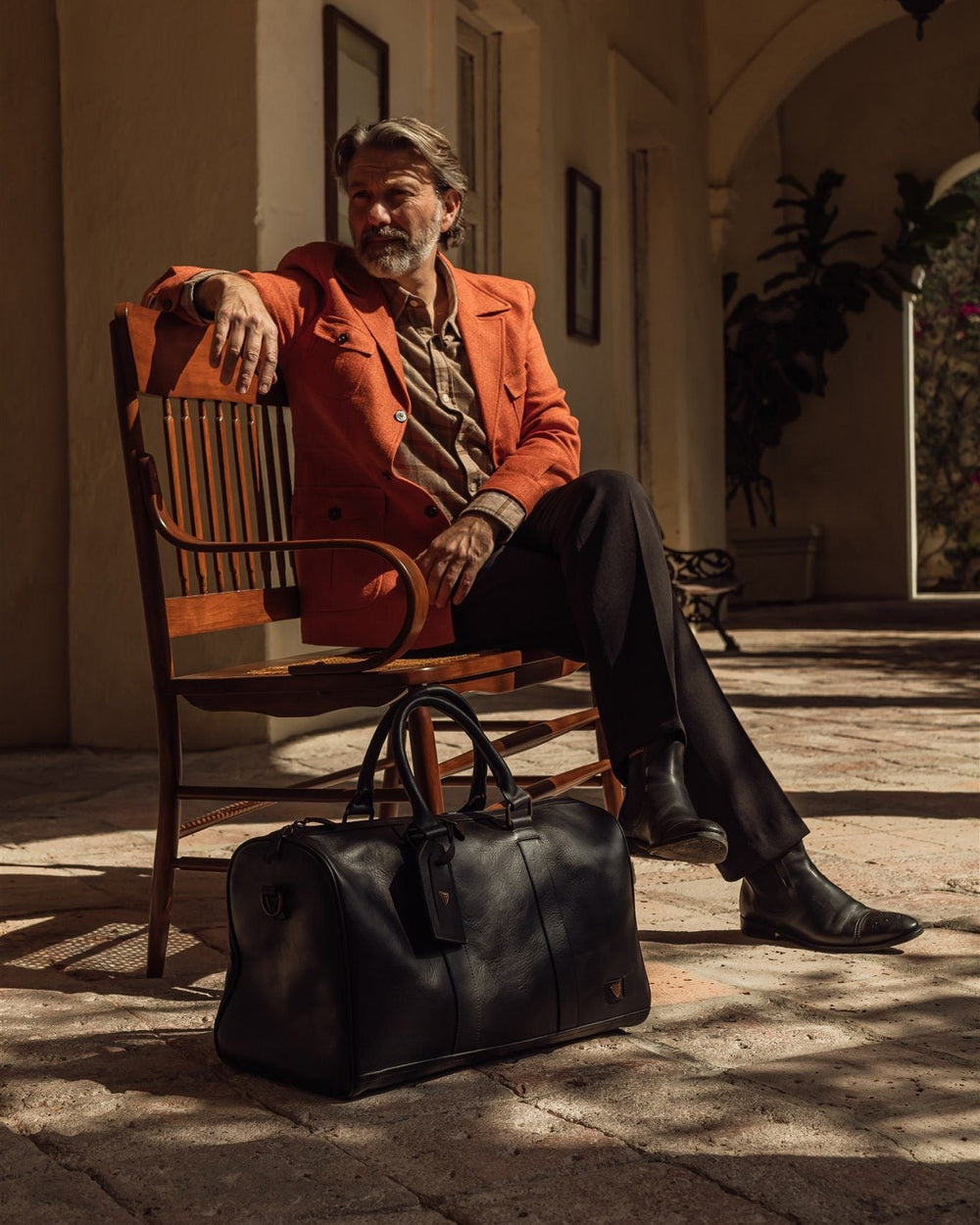 Man in a rust overcoat seated in a sunlit Mexican arcade, a black leather duffle at his feet