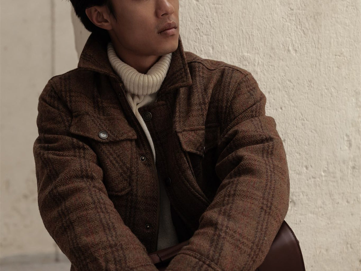 Man in a brown plaid wool jacket and cream turtleneck against a white wall