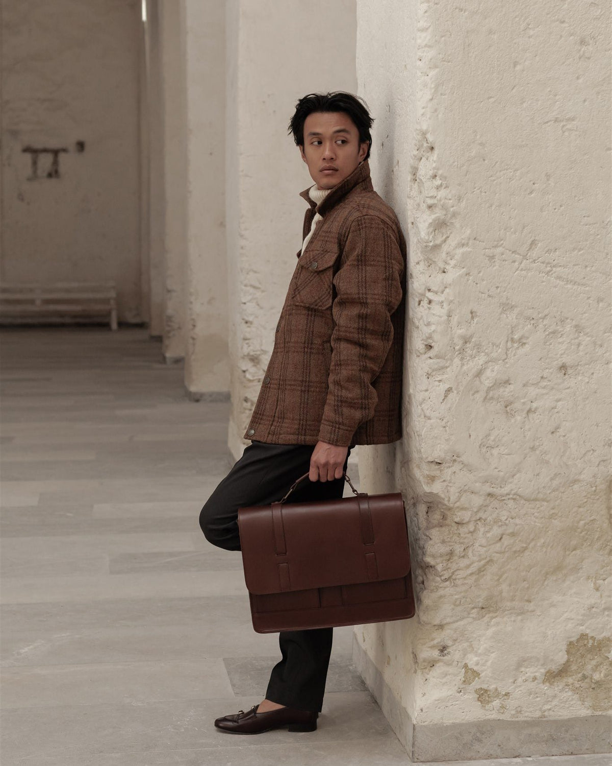 Man in a brown plaid wool jacket leaning on a white arcade pillar, holding a brown leather attaché