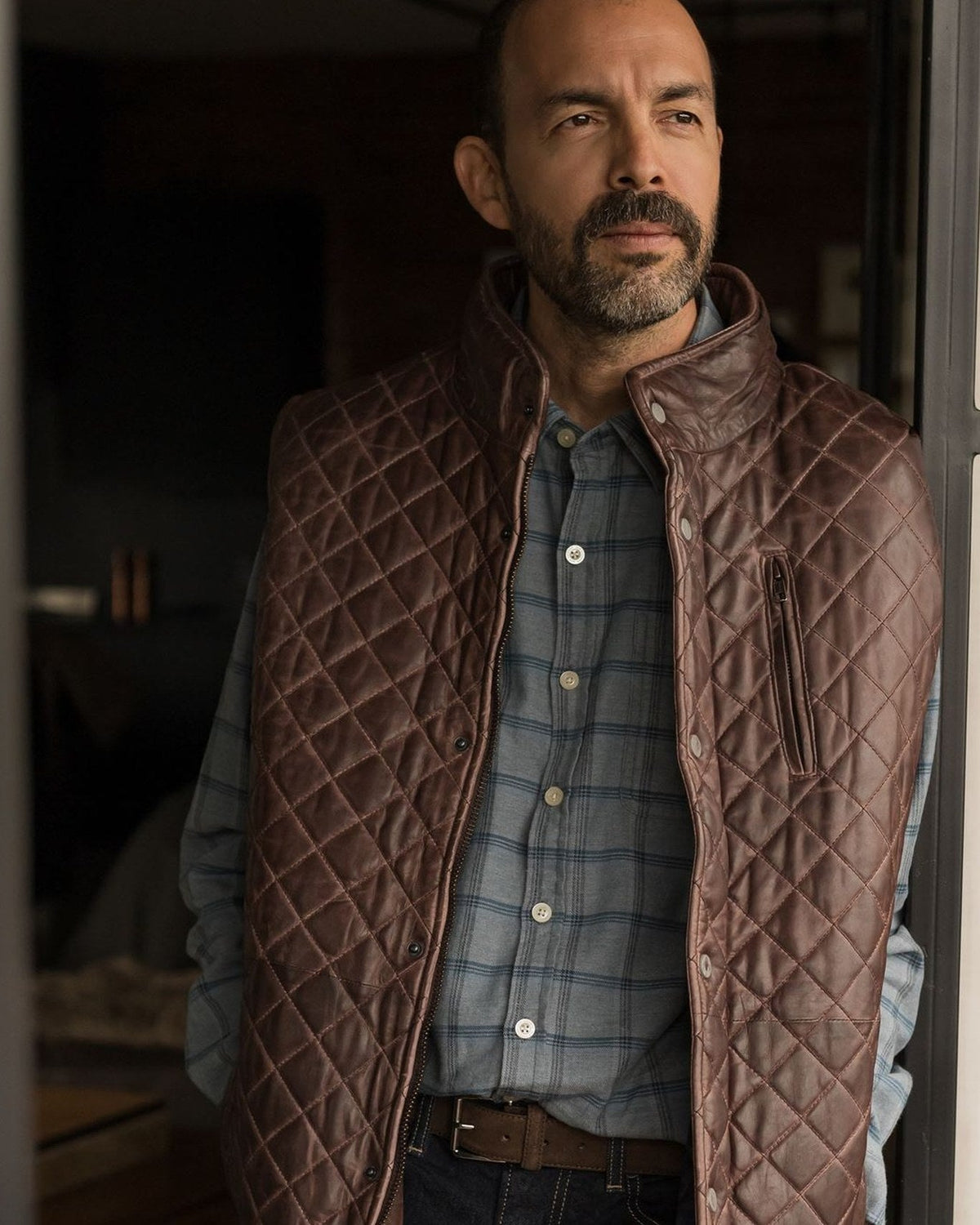Man in the Windsor Cotton Shirt in Sky Pane under the Highlands Quilted Leather Vest in Mahogany Brown, standing in a doorway