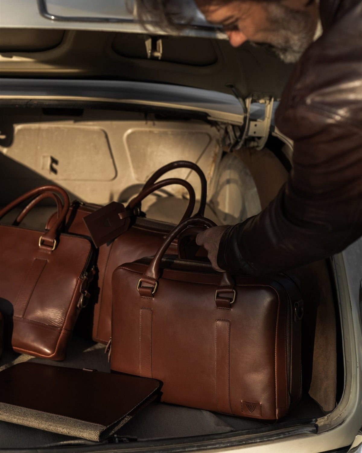 Man in a leather jacket loading a duffle, briefcase and attaché into the trunk of a vintage car