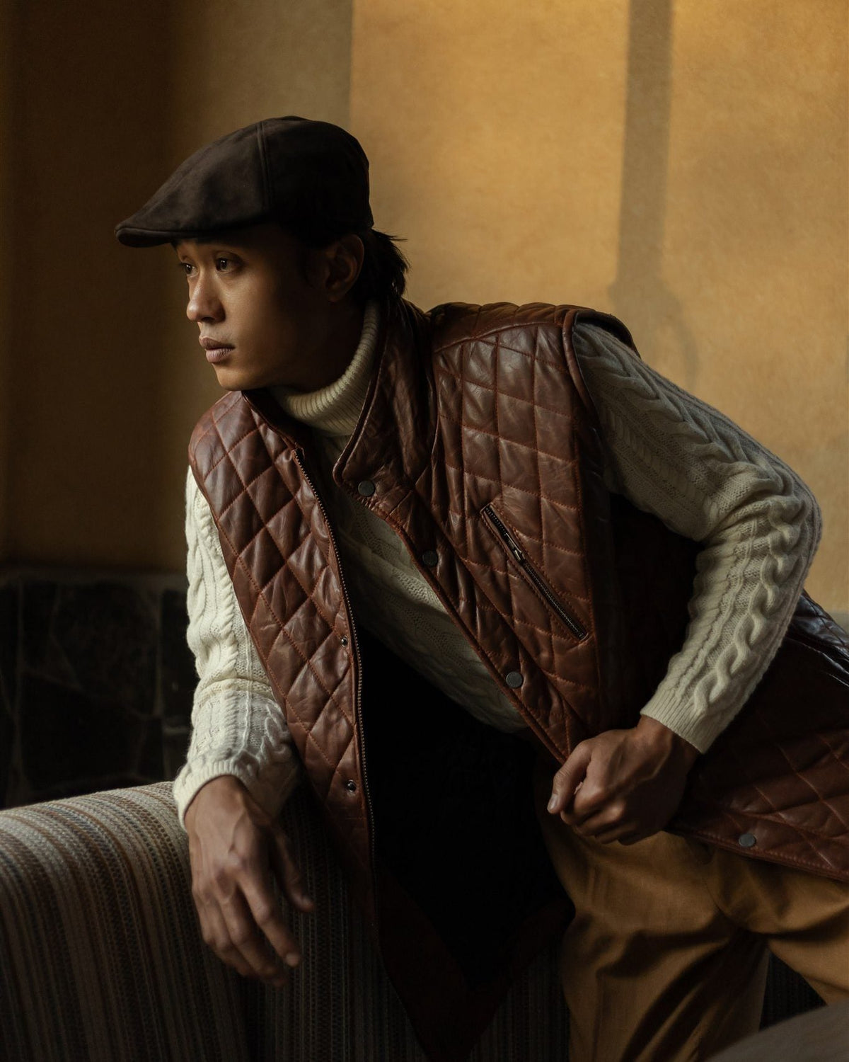 Man in a mahogany quilted leather vest, cable knit and flat cap, seated by a window