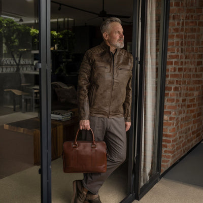 Man in a leather jacket standing in a doorway with the Jefferson Leather Attaché Briefcase in Elderwood Brown.