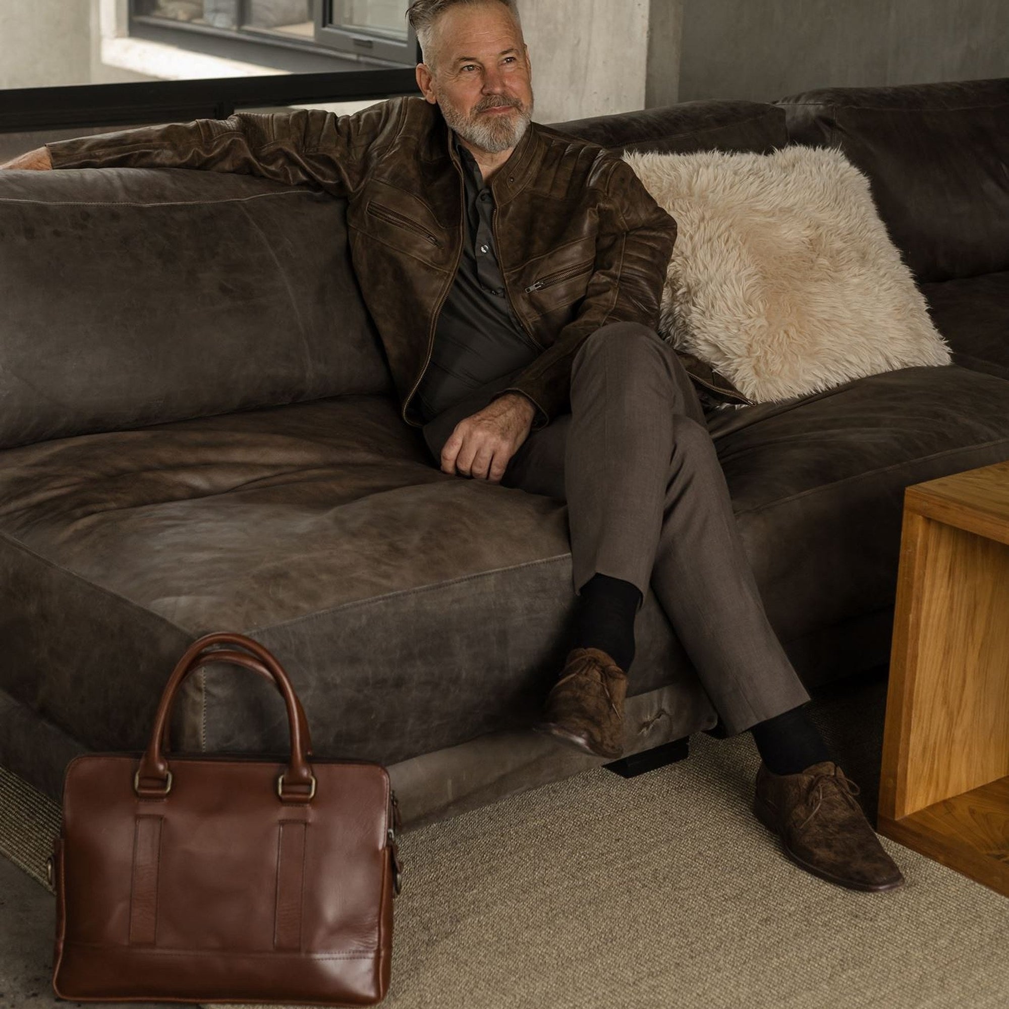 Man seated on a leather sofa with the Jefferson Leather Attaché Briefcase in Elderwood Brown at his feet.