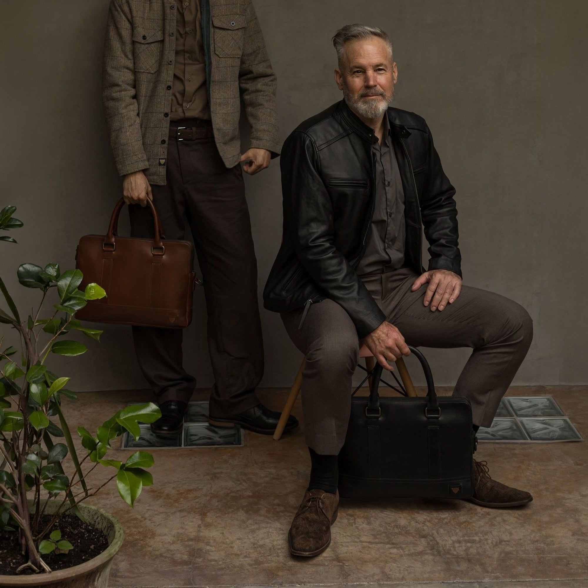 Seated man with the Jefferson Leather Attaché Briefcase in Vintage Black at his feet, beside a man holding the Elderwood Brown.