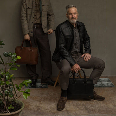 Seated man with the Jefferson Leather Attaché Briefcase in Vintage Black at his feet, beside a man holding the Elderwood Brown.