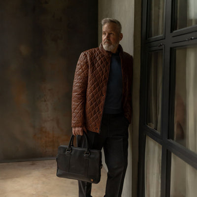 Man in a quilted leather jacket by a window, carrying the Jefferson Leather Attaché Briefcase in Vintage Black.