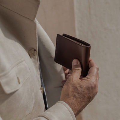 Hand holding the Jefferson Leather Bifold Wallet in Elderwood Brown against a cream linen jacket.