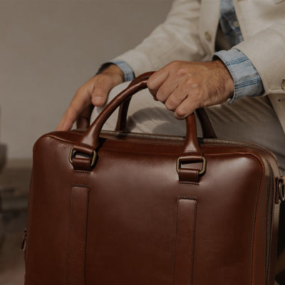Hand on the top handles of the Jefferson Leather Briefcase in Elderwood Brown.