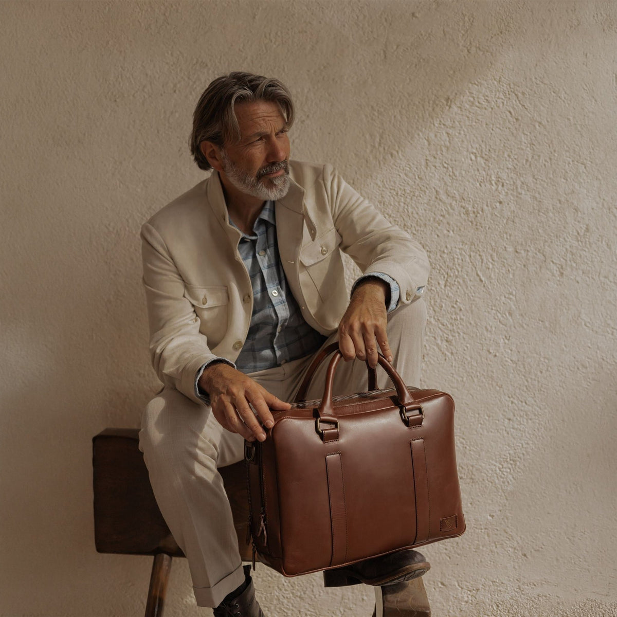 Man in a cream linen jacket seated against a sunlit wall, holding the Jefferson Leather Briefcase in Elderwood Brown.