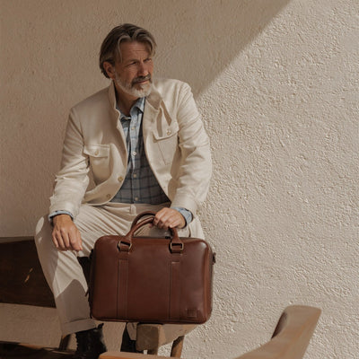 Man in a cream linen jacket seated with the Jefferson Leather Briefcase in Elderwood Brown.