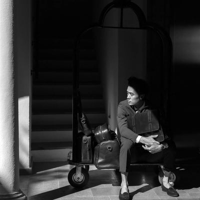 Man seated on a hotel luggage cart loaded with the Jefferson leather collection in Vintage Black, in black and white.