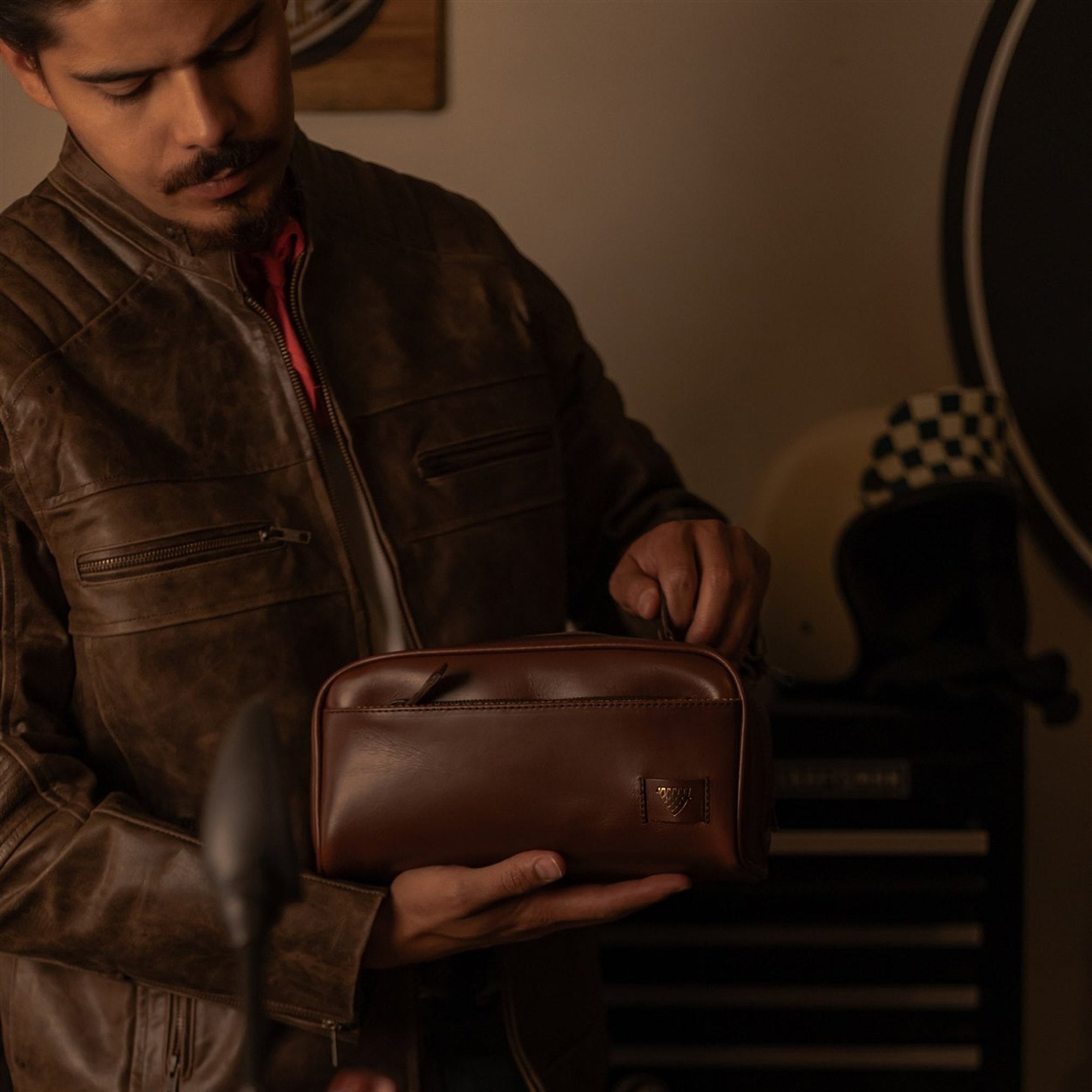 Man in a leather jacket packing the Jefferson Leather Dopp Kit in Elderwood Brown in a motorcycle garage.