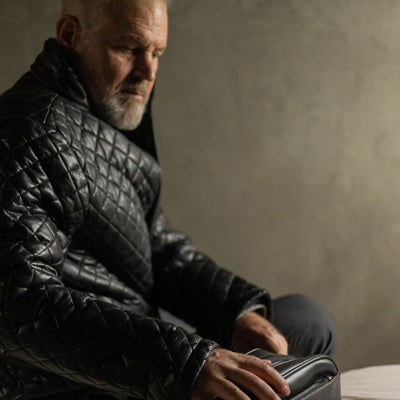 Man in a black quilted jacket seated on a bed with the Jefferson Leather Dopp Kit in Vintage Black.