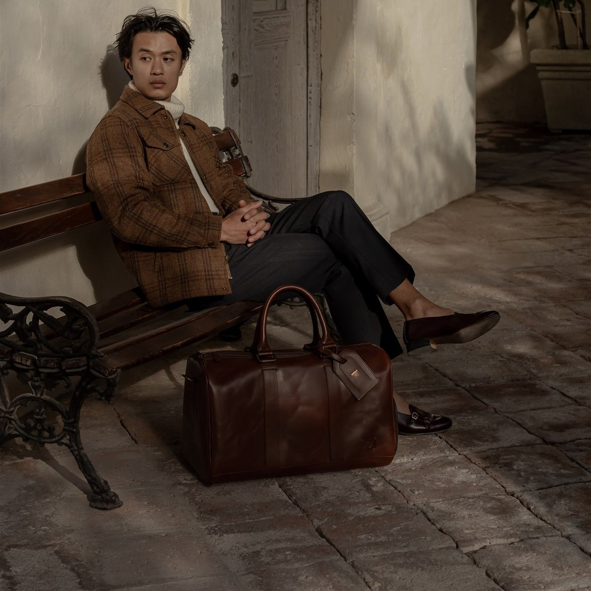 Man in a brown wool jacket seated on a bench beside the Jefferson Leather Duffle in Elderwood Brown