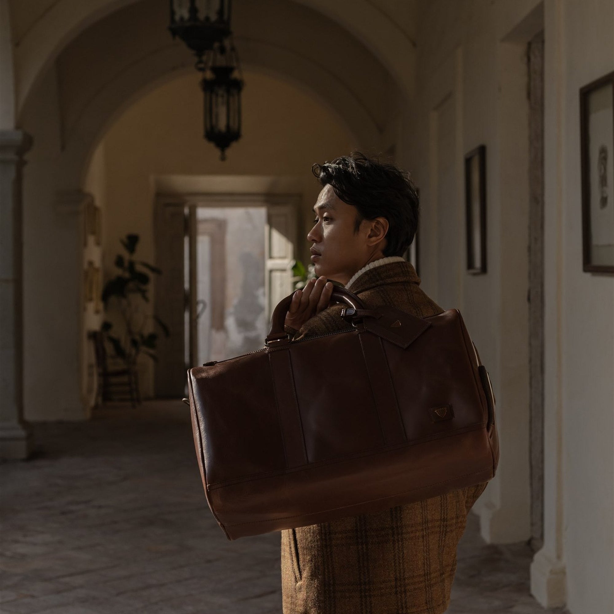 Man holding the Jefferson Leather Duffle in Elderwood Brown in an arched hallway.