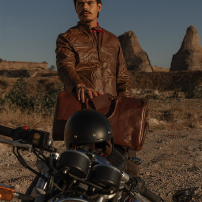 Man resting a hand on the Jefferson Leather Duffle in Elderwood Brown behind a motorcycle in the desert.