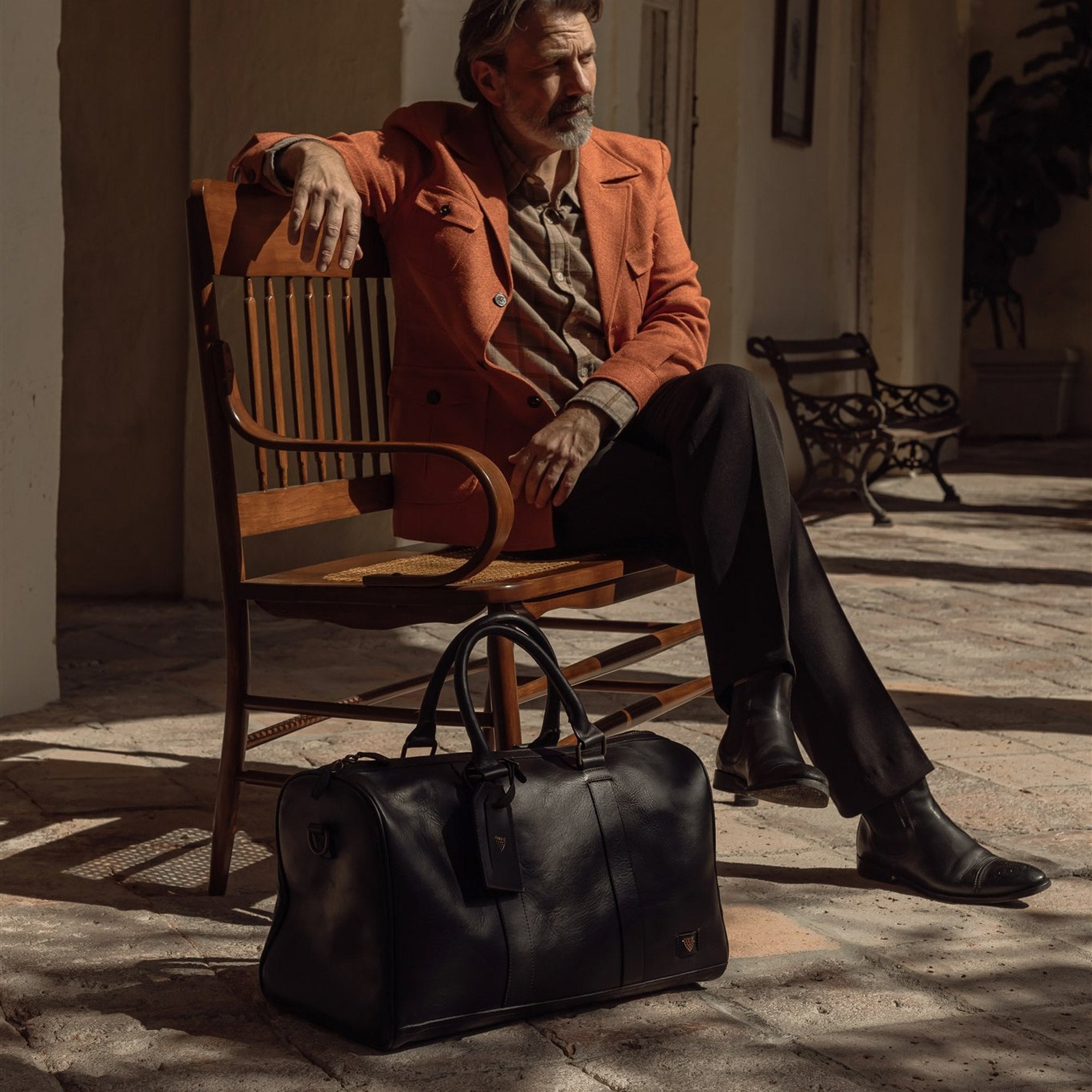 Man seated on a bench in an arcade with the Jefferson Leather Duffle in Vintage Black at his feet.