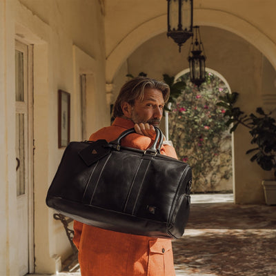 Man in an orange jacket carrying the Jefferson Leather Duffle in Vintage Black under stone arches.