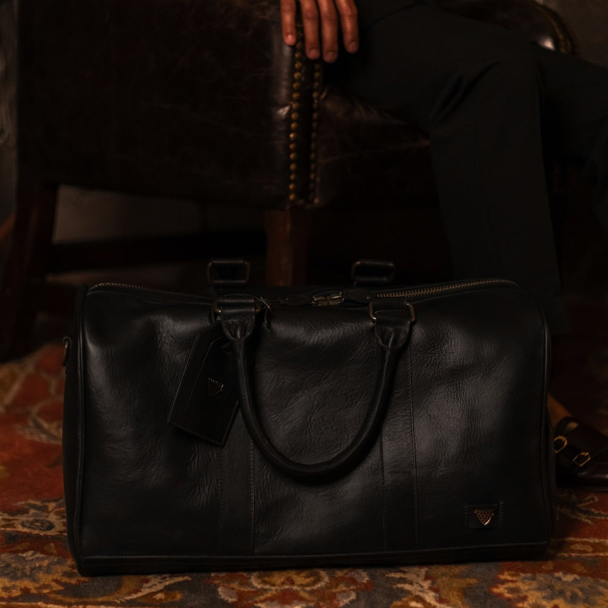 Jefferson Leather Duffle in Vintage Black on a patterned rug beside a leather chair.