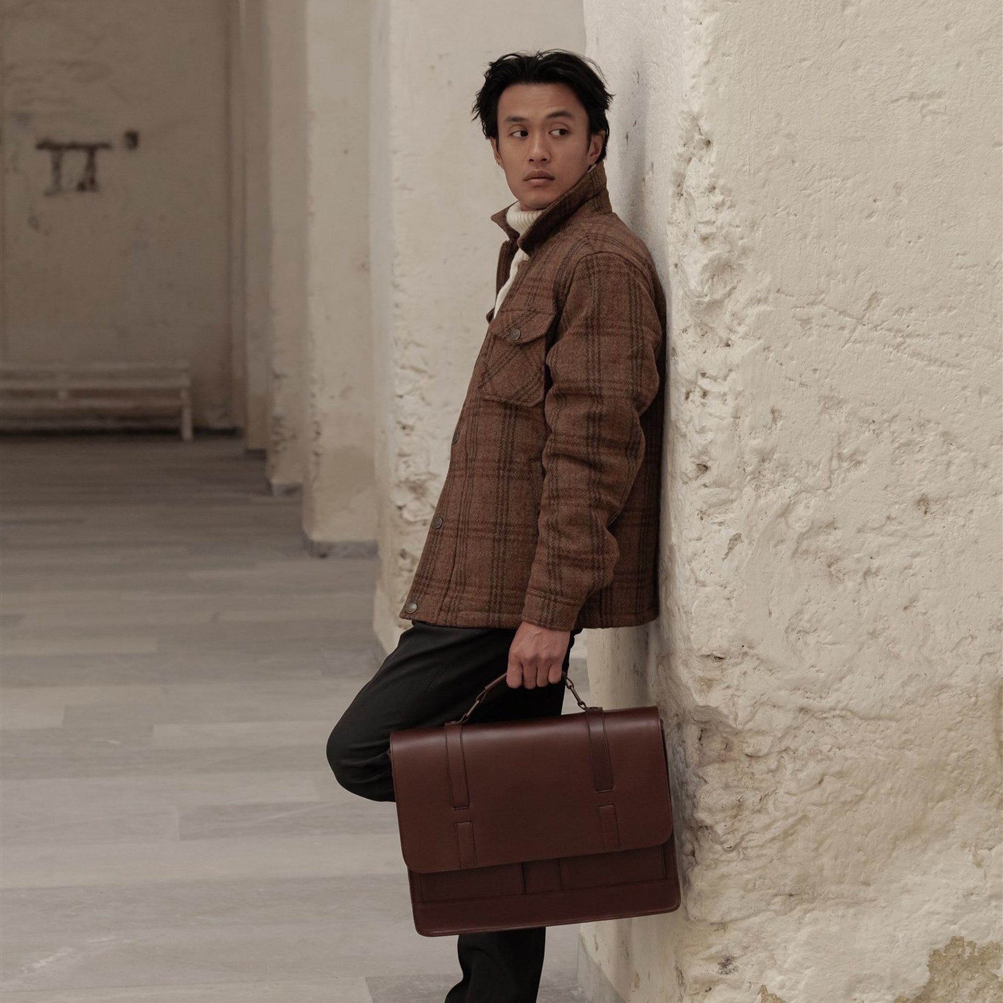 Man leaning on a column with the Jefferson Leather Messenger Bag in Elderwood Brown.