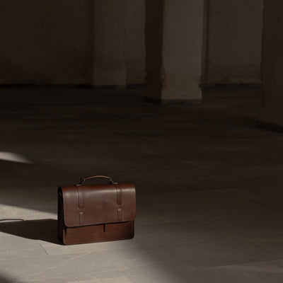The Jefferson Leather Messenger Bag in Elderwood Brown on a stone floor in a pool of light.