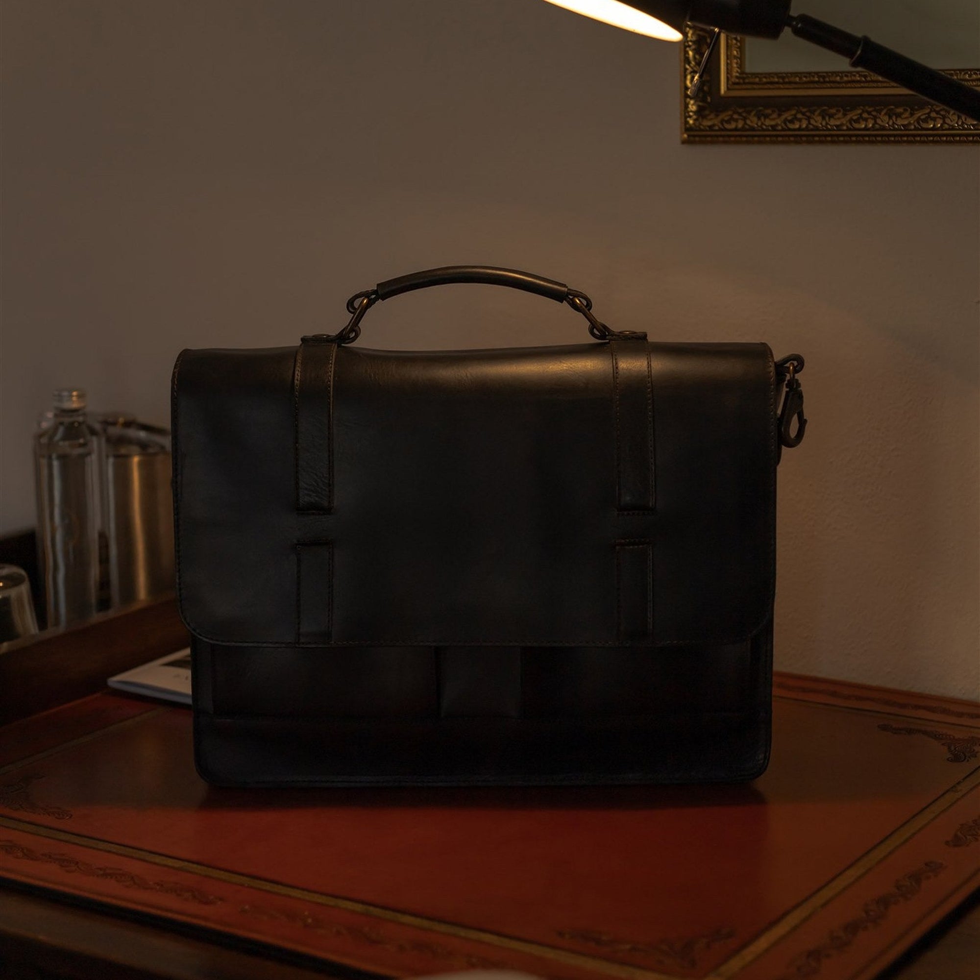 The Jefferson Leather Messenger Bag in Vintage Black on a desk under a reading lamp.