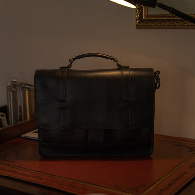 The Jefferson Leather Messenger Bag in Vintage Black on a desk under a reading lamp.